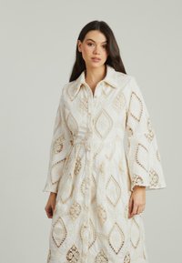 Abito bianco con motivi a uncinetto a forma di diamante, maniche a campana e chiusura frontale con bottoni. Presenta dettagli in pizzo intricati e una trama morbida.