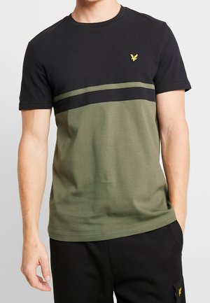 Mężczyzna ubrany w czarny i oliwkowo-zielony t-shirt z krótkim rękawem z małym żółtym logo ptaka na piersi.