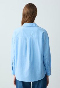 Camicia over-size azzurro chiaro con colletto, maniche lunghe e polsini con bottoni. Presenta una piega sul retro e un orlo curvo, realizzata in tessuto morbido.