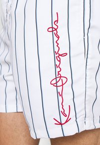Shorts de bain blancs avec des rayures verticales bleues et noires. Présente un logo rouge brodé sur le côté. Tissu lisse et léger.