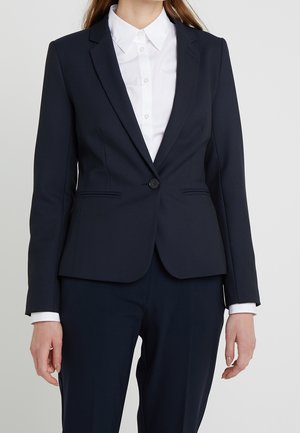 Woman wearing a tailored navy blazer buttoned once over a white collared shirt with matching navy trousers, standing against a plain background.