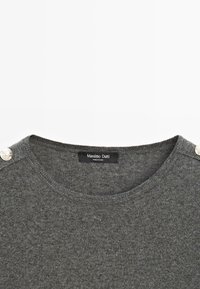 Maglione grigio lavorato a maglia con scollo rotondo, caratterizzato da due bottoni decorativi argentati sulla spalla sinistra e un'etichetta del marchio all'interno.