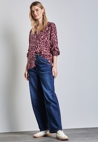 Bordeaux gekleurde blouse met patroon en lange mouwen, gecombineerd met wijde, donkere spijkerbroek en witte sneakers. Gelaagde stof en subtiele ontwerpdetails.