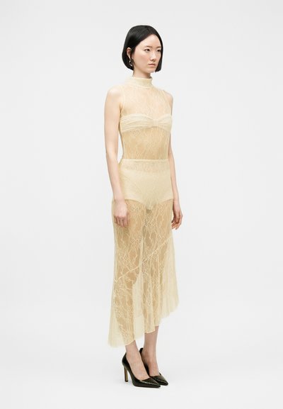 Γυναίκα με κοντά μαύρα μαλλιά φοράει διαφανές μπεζ δαντελένιο φόρεμα με ψηλό λαιμό, τοπ μπαντό και μαύρα μυτερά ψηλοτάκουνα, στέκεται σε λευκό φόντο.