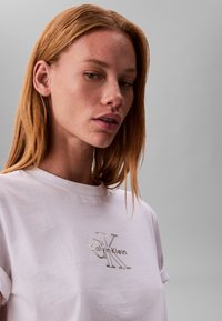 Junge Frau mit langen roten Haaren, die ein weißes Calvin Klein T-Shirt trägt und leicht seitlich vor einem schlichten grauen Hintergrund schaut.