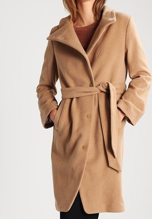 Femme portant un manteau en laine couleur camel ceinturé avec des boutons-pression et les mains dans les poches, sur un haut marron et un pantalon noir.