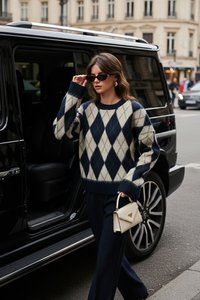 Vrouw met argyle trui en zonnebril stapt uit een zwarte SUV en houdt een kleine witte handtas vast op een stadsstraat.