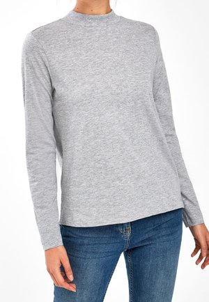 Persoon draagt een lichtgrijze top met lange mouwen en een hoge halslijn, gecombineerd met blauwe spijkerbroek, tegen een witte achtergrond.