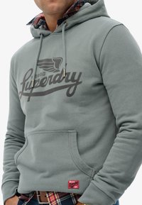 Grijze capuchontrui met een voorzak, voorzien van een groot zwart "Superdry" logo, trekkoorden en een rood merkembleem aan de zoom.