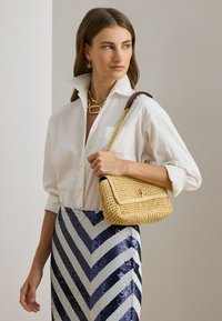 Γυναίκα που φοράει λευκό πουκάμισο με κουμπιά και μπλε-άσπρη ριγέ φούστα με παγιέτες, κρατώντας ένα πλεκτό μπεζ τσαντάκι με χρυσή αλυσίδα.