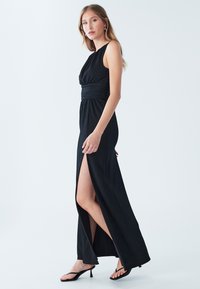 Schwarzes ärmelloses Maxikleid mit geraffter Taille, hohem Schlitz bis zum Oberschenkel und glatter Textur, kombiniert mit schwarzen, lässigen Sandalen mit hohen Absätzen.