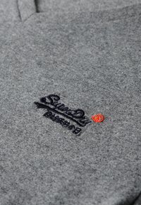 Grå bomulds polo skjorte med V-hals design. Har broderet logo i mørkeblå med orange accent. Blød tekstur, afslappet stil.