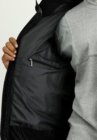 Interior de chaqueta negra que muestra un bolsillo con cremallera, llevada sobre un sudadera gris con textura y un sutil patrón de espiga.