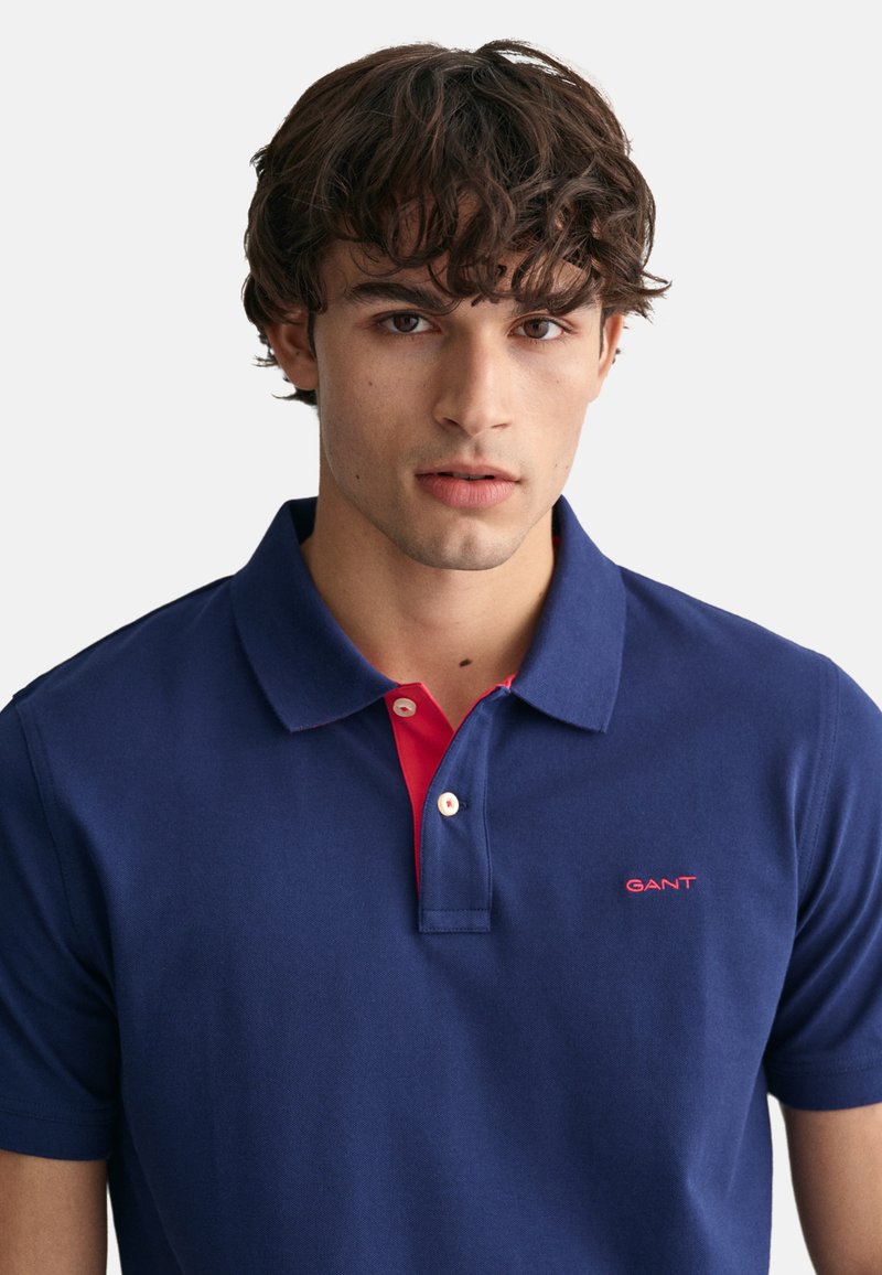 Junger Mann mit lockigem braunem Haar, der ein dunkelblaues Poloshirt mit einer roten Inneneinfassung und einem roten GANT-Logo auf der Brust trägt.