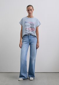 Eine Frau steht da und trägt ein hellblaues Vintage-Grafik-T-Shirt, eine weit geschnittene hellblaue Jeans und weiße Sneaker vor einem einfarbigen Hintergrund.