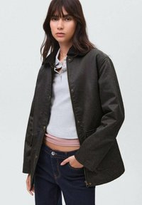 Rjava vodoodporna jakna z teksturirano površino, ki ima ovratnik in pritisne gumbke, nosi se preko svetlo sive majice in temnih kavbojk.