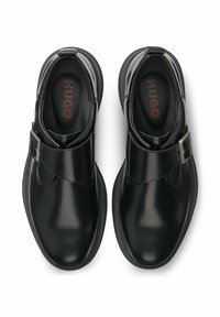 Scarpe in pelle verniciata nera con punta arrotondata, cinturino regolabile con fibbia e finitura liscia. Interno con logo "HUGO" rosso.