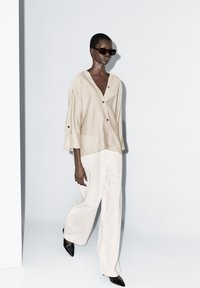 Beige oversize košile z lněného materiálu s hlubokým véčkovým výstřihem a knoflíky, v kombinaci s bílými kalhotami s širokými nohavicemi a špičatými černými botami. Sluneční brýle dotvářejí vzhled.