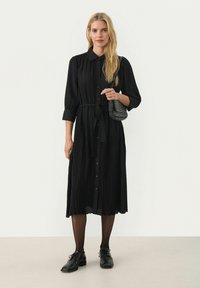 Abito nero plissettato con scollatura a colletto, chiusura a bottoni davanti e cintura in vita; abbinato a una borsa texture e scarpe nere.