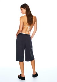 Schwarze Baumwollshorts mit lockerem Schnitt und seitlichen Taschen, kombiniert mit einem weißen BikinOberteil mit Schnürung, Modell trägt Plateausandalen.