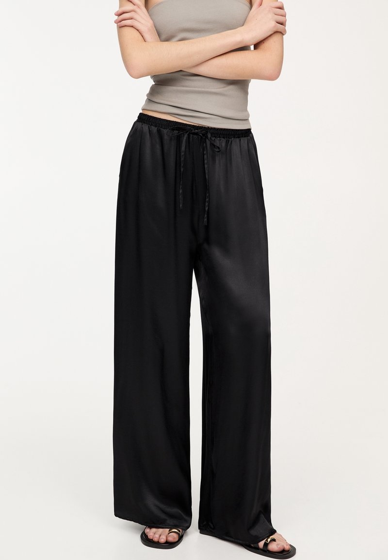 Personne portant un pantalon large en satin noir avec taille à cordon, un haut sans manches beige et des sandales noires sur un fond uni.