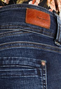 Ciemnoniebieskie dżinsy o teksturowanym wykończeniu, z brązową skórzaną metką, miedziowanymi elementami oraz widocznymi detalami szwów.