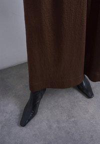 Άτομο που φορά καφέ υφασμάτινο παντελόνι με φαρδιά ποδάρια και μαύρα δερμάτινα μποτάκια με τετράγωνη μύτη, όρθιο σε γκρι τσιμεντένιο πάτωμα.