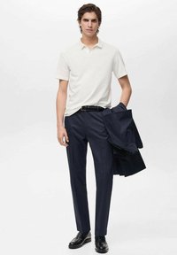 Witte polo, marineblauwe dresspantalon, zwarte riem, zwarte instappers; houdt een marineblauwe jas vast. Gladde stof, slimfit ontwerp, strakke lijnen.