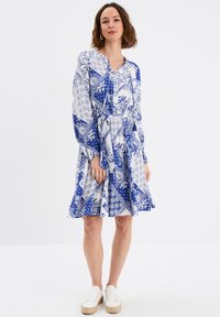 Vestido azul y blanco con estampado, de mangas largas, con un lazo en la cintura y una falda acampanada. La tela presenta diseños y texturas intrincados.