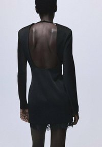 Vestido negro con mangas largas y un profundo escote en la espalda. Presenta un delicado borde de encaje en el dobladillo. Tela suave, diseño ajustado.