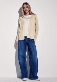 Beige öppen kofta över en vit tryckt t-shirt, i samverkan med vida blåjeans. Slät textur och avslappnad design. Förstärkt med en logotyp.