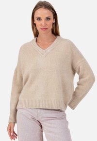 Beiger V-Ausschnitt-Pullover aus weichem, strukturiertem Strick mit heruntergesetzten Schultern und gerippten Bündchen, kombiniert mit hellrosa Hosen.