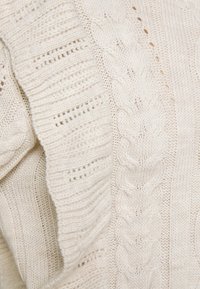 Gros plan sur un tissu tricoté couleur crème montrant des motifs torsadés et côtelés avec des points texturés.