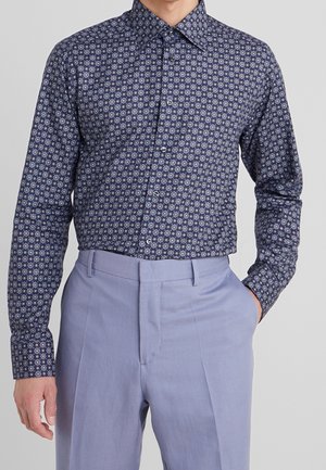 Mand iført langærmet skjorte med blå blomsterprint, stoppet ned i lyseblå skræddersyede bukser, med den ene hånd i lommen.