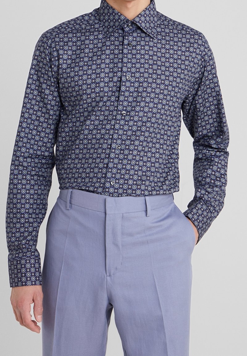 Homme portant une chemise boutonnée à manches longues avec un motif floral bleu rentrée dans un pantalon sur-mesure bleu clair, une main dans la poche.