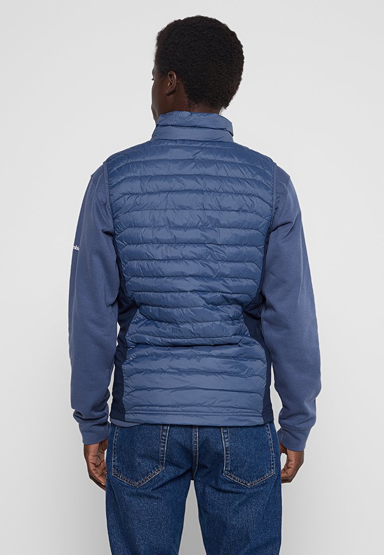 Uomo che indossa un gilet imbottito blu sopra una camicia a maniche lunghe blu e jeans blu, in piedi rivolto di spalle contro uno sfondo semplice.