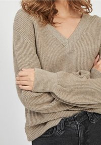 Pull beige à col en V avec une texture côtelée et des manches larges, associé à un jean gris foncé. Le tissu semble doux et confortable.