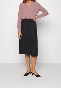 Camicia a maniche lunghe button-up di un delicato rosa chiaro abbinata a una gonna midi nera plissettata. Modella che indossa tacchi neri a punta. Design semplice ed elegante.