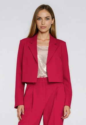 Krótka marynarka w jasnoróżowym materiale z wciętymi klapami, zestawiona z beżowym topem wyszywanym cekinami. Dopasowany krój z długimi rękawami i zapięciem na guziki.