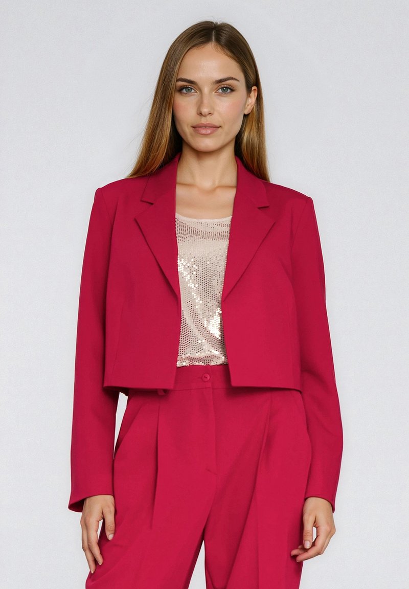 Krótka marynarka w jasnoróżowym materiale z wciętymi klapami, zestawiona z beżowym topem wyszywanym cekinami. Dopasowany krój z długimi rękawami i zapięciem na guziki.