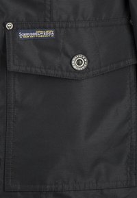 Chaqueta de tela negra con un bolsillo en el pecho que tiene una etiqueta azul y un botón a presión plateado, textura suave y diseño sencillo.