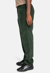 Green corduroy trousers with a straight leg design, featuring subtle texture and side pockets, paired with black athletic sneakers.
