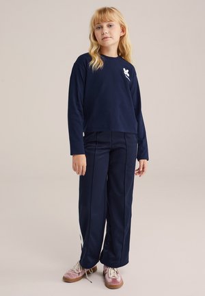 Mörkblå långärmad skjorta med vit grafik, kombinerat med mörkblå byxor med sidostråkar. Modellen har på sig rosa skor med vita snören.