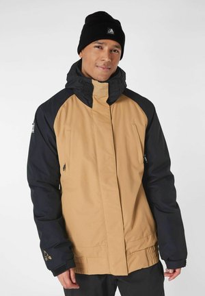 Waterdichte jas met een tan kleurig lijf en marineblauwe mouwen, verstelbare capuchon, ritssluitingenzakken en elastische manchetten voor extra comfort.