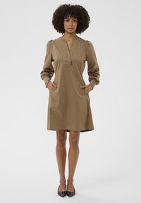 Göndör hajú nő, aki khaki színű, hosszúujjú, térdig érő ruhát visel zsebekkel, egyenesen áll egy sima fehér háttér előtt.