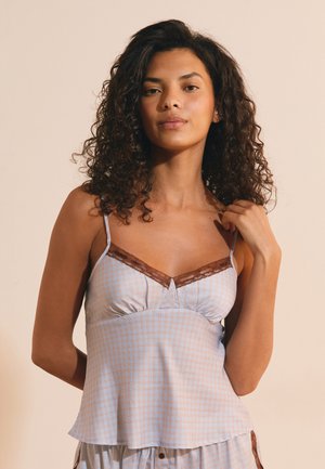 Vrouw met krullend haar die een mouwloos geruit topje met kanten rand draagt, staand tegen een effen beige achtergrond.