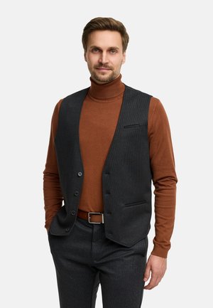 Man met kort bruin haar draagt een bruine coltrui, donkergrijs vest, bijpassende broek en een bruine riem, staat en glimlacht.