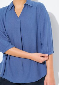 Blusa blu a maniche corte con colletto e vestibilità rilassata. Il tessuto sembra morbido con una sottile texture e polsini delle maniche arrotolati.