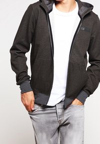 Felpa con zip di colore grigio scuro, con una fodera interna grigio chiaro a contrasto, polsini a costine e tasche frontali. Presenta un piccolo logo sul petto.