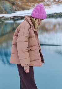 Hnedá prešívaná bunda s vysokým golierom a prednými vreckami, v kombinácii s tmavými nohavicami. Celý vzhľad dopĺňa jasnoružová pletená čiapka.